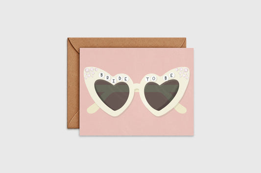 Bride To Be engagement card with white heart-shaped sunglasses on blush pink, with kraft envelope