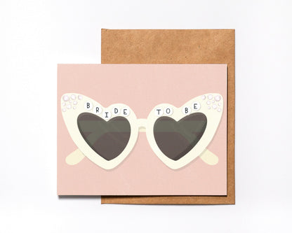 Bride To Be engagement card with white heart-shaped sunglasses on blush pink, with kraft envelope