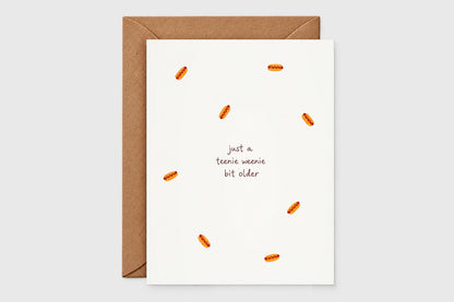 Teenie weenie bit older birthday card with scattered hot dog illustrations on a white card, with kraft envelope